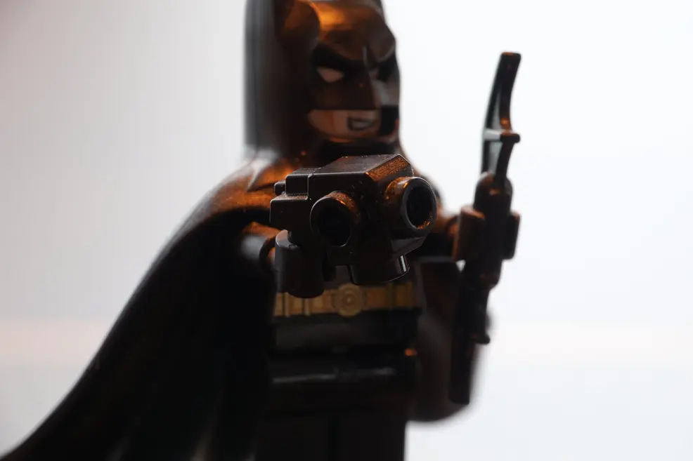 Close-up of a LEGO Batman minifigure with a black cape and a grin, aiming a black grappling gun at the camera against a bright, blown-out background