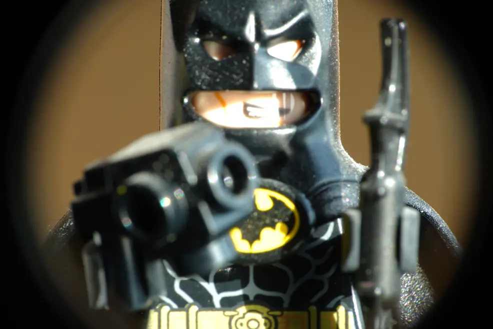 Close-up of a LEGO Batman minifigure in sunlight, pointing a blaster towards the camera, framed by a dark round vignette