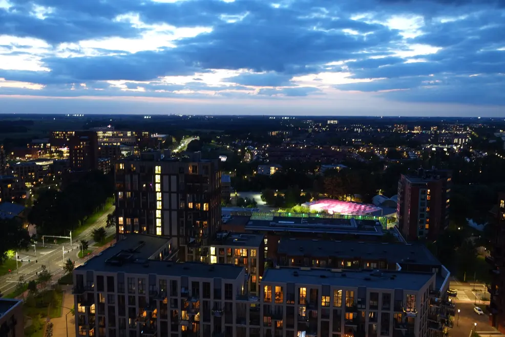 Dusk over Den Bosch seen from a high floor: blue clouds with a pink glow on the horizon above residential towers with lit windows, and a greenhouse lit pink and green among the trees