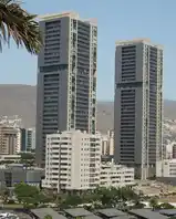 Two tall grey residential towers on Tenerife, side by side against a blue sky, with lower buildings, palm trees and a parking lot in front