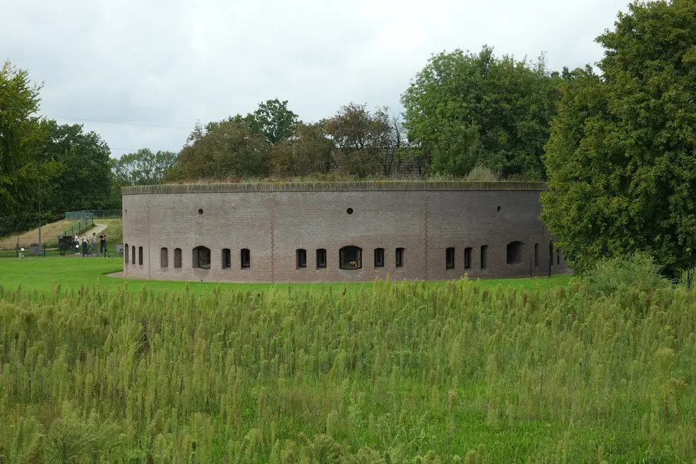 A round brick fort with small arched windows and a grass-covered roof, seen across a field of tall green grass, with trees behind it and a few visitors on the path to its left under an overcast sky