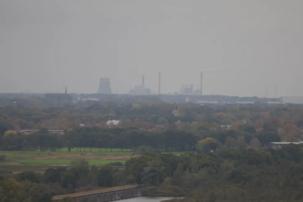 A hazy autumn landscape seen through a long lens: forest in orange, yellow and green and a strip of meadow in front, and on the skyline the power plant of Geertruidenberg, with a cooling tower and chimneys, one with a thin plume of smoke, under a flat grey sky