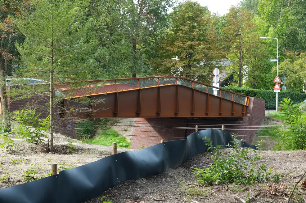 A rust-coloured steel bridge with a brick abutment and a mesh railing, seen from the side across a construction fence, with a car just visible behind the railing on the left, and road signs on the right: two round prohibition signs, a priority sign with arrows, and a triangular warning sign, among green trees