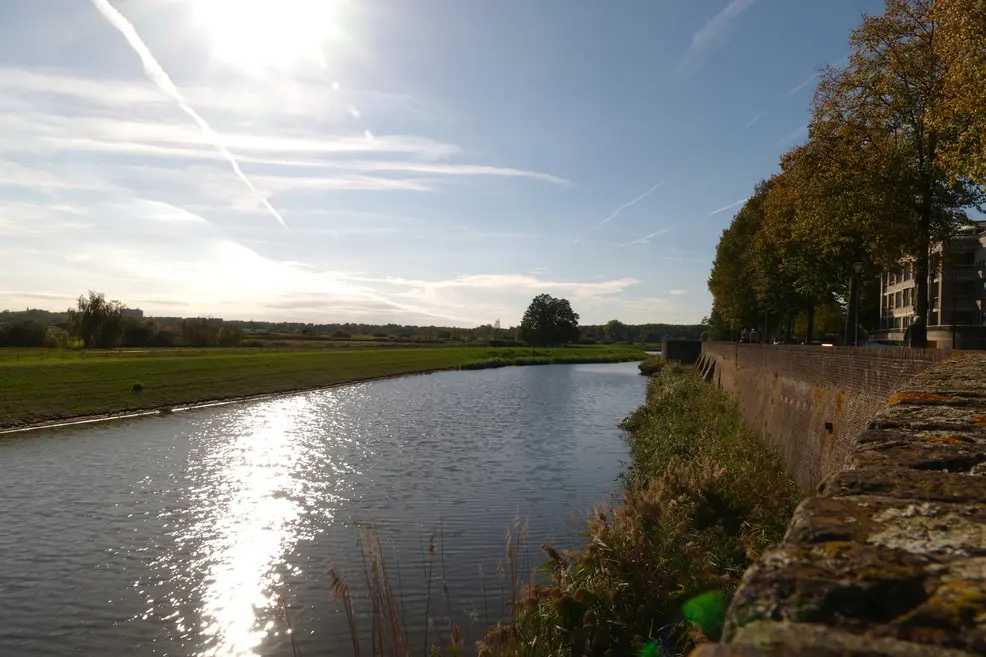 Low afternoon sun reflecting on a moat that runs along an old brick city wall, with autumn trees, a green meadow and a sky full of contrails