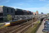A yellow and blue train blurred into a streak by a long exposure as it passes under overhead wires, with sharp office buildings and a parking lot around it