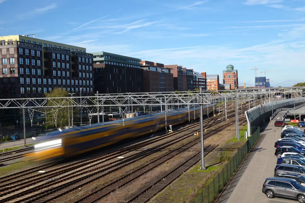 A yellow and blue train blurred into a streak by a long exposure as it passes under overhead wires, with sharp office buildings and a parking lot around it