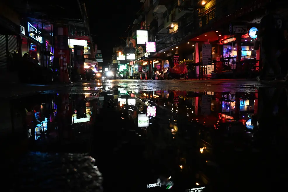 A dark street at night in Pattaya, with neon and lit signs in pink, blue, red and white reflected in a large puddle in the foreground