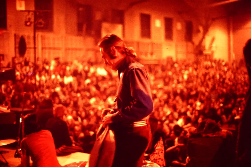 Timothy Leary in a dark jacket, seen from the side on a stage in red-orange light, in front of a large crowd